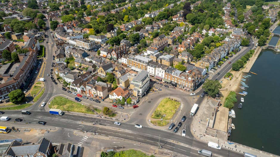 An aerial view of a small town showing a mix of residential and commercial buildings with various roof styles and colors, surrounded by lush green trees and open spaces. The streets are bustling with 